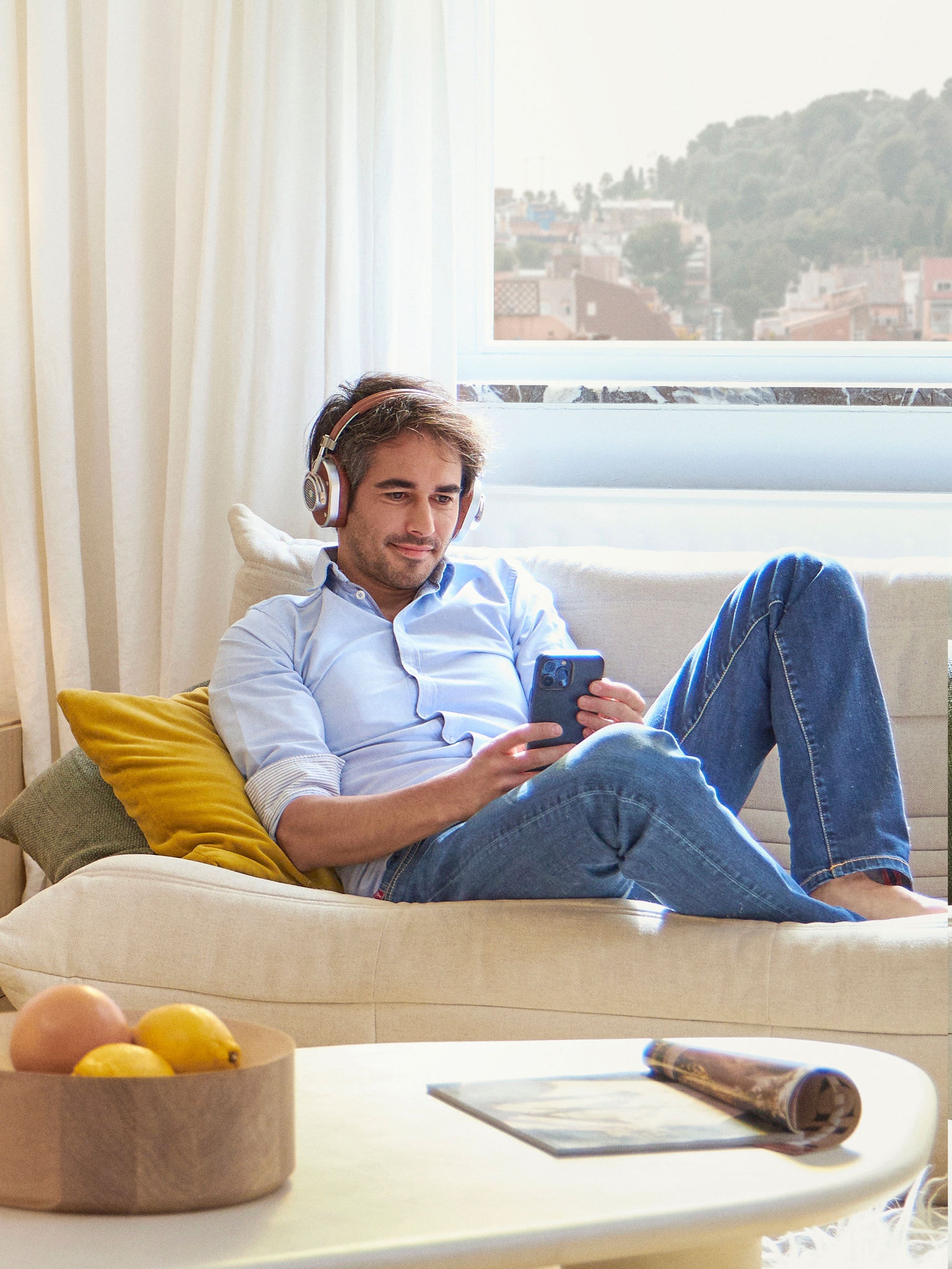 Човек със слушалки, излежаващ се на дивана си, докато сърфира на мобилния си телефон